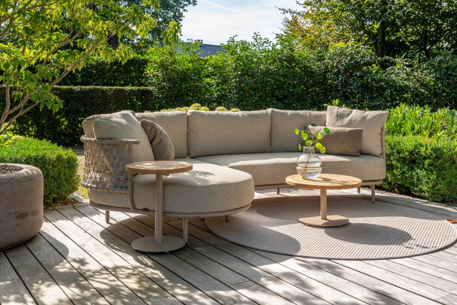 Moderne beige loungebank op een houten terras, met twee ronde bijzettafels en veel groen in de tuin eromheen