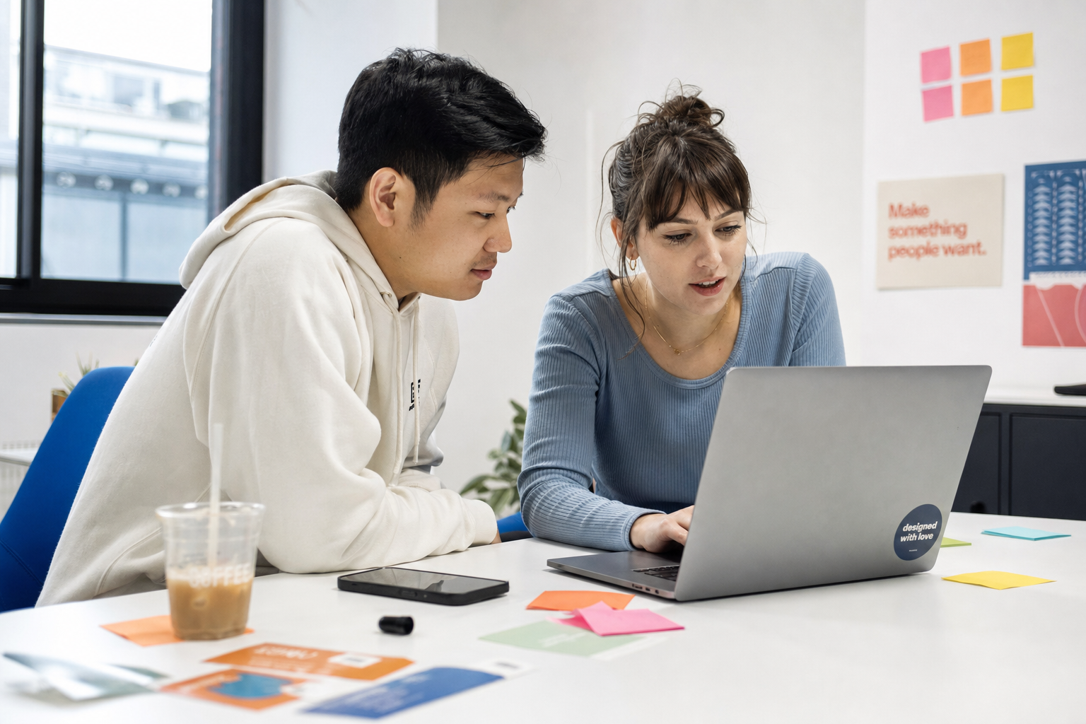 Twee collega's werken samen aan een laptop aan een bureau met notities en koffie.